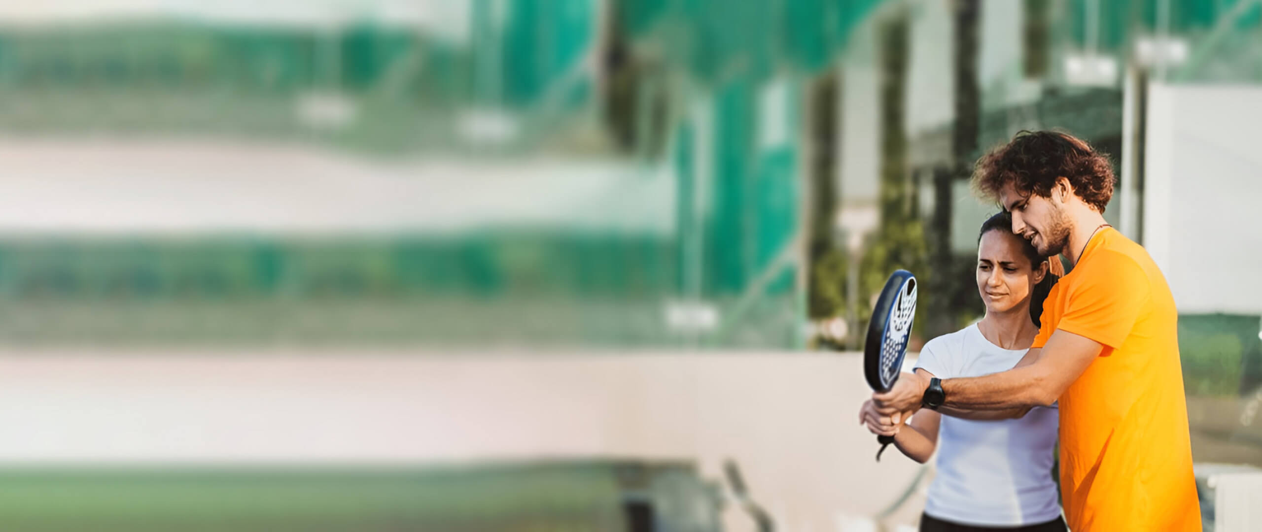 A male padel tennis coach demonstrating proper racket technique to a female student on an outdoor court, perfect for a guide on how to play padel.